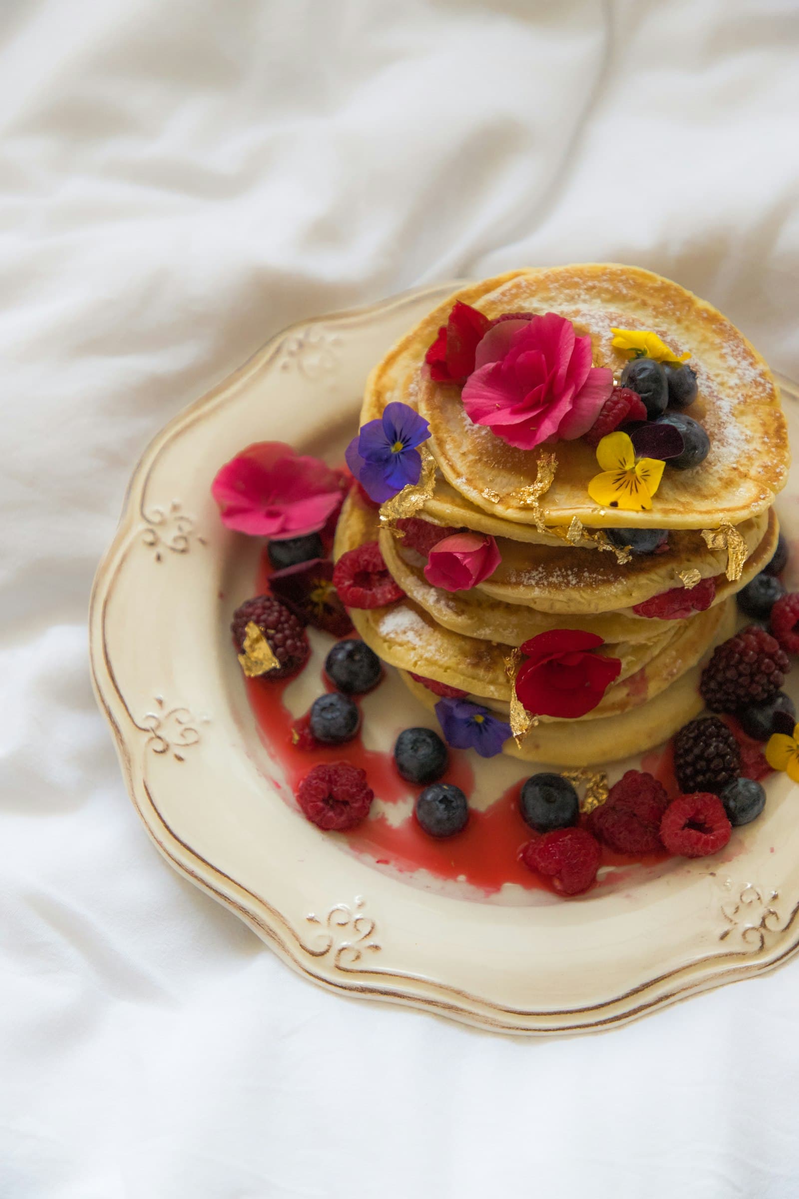 A stack of pancakes crowned with edible flowers and fresh berries on an ornate plate in soft daylight