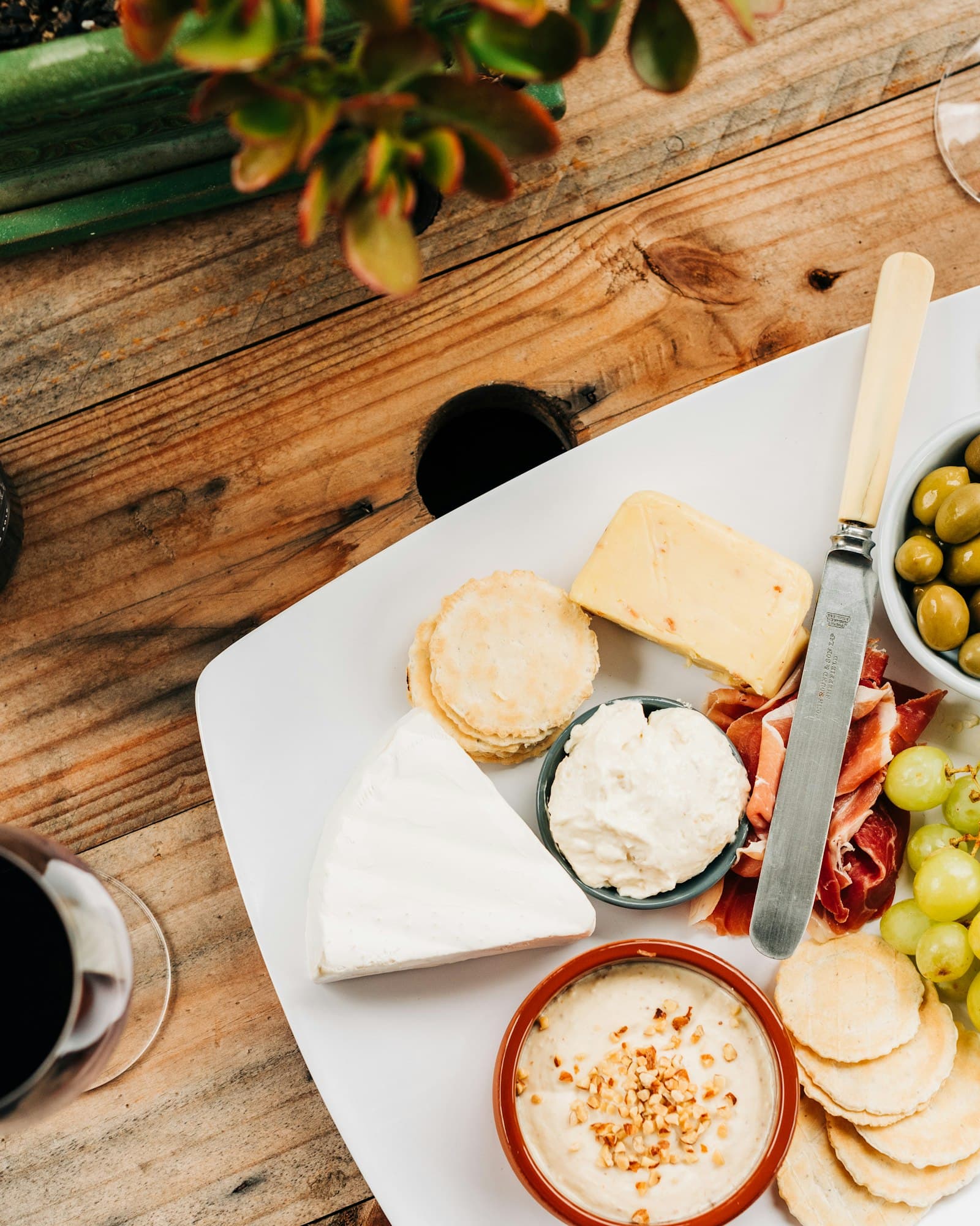 A charcuterie and cheese board with prosciutto, olives, and grapes beside a glass of red wine on a warm wood table